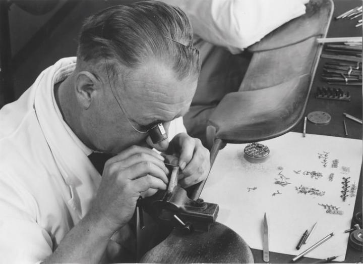 时值1963年，Bienne工坊里的专业工匠正以放大镜仔细制作模具，而台面上精致小巧的零件正待装载，工匠投入高度集中力才能成功装配好一枚机芯