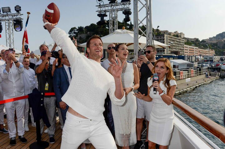 知名四分卫Tom Brady在游艇甲板边，朝著小艇上的Daniel Ricciardo掷美式足球，完美达成任务