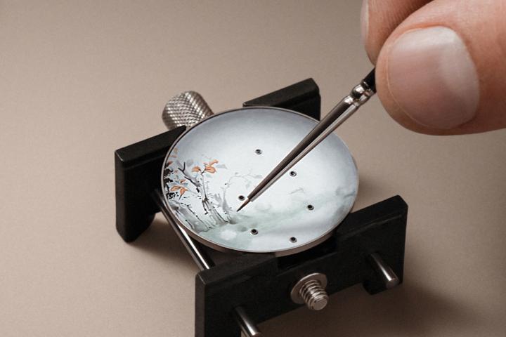 表盘背景的刻画结合了两种精妙的珐琅技艺：先以多色渐变的大明火珐琅工艺，在表盘上呈现从中央向边缘由浅至深的色调过渡；随后运用微绘珐琅技法勾勒出繁茂的植物纹样。