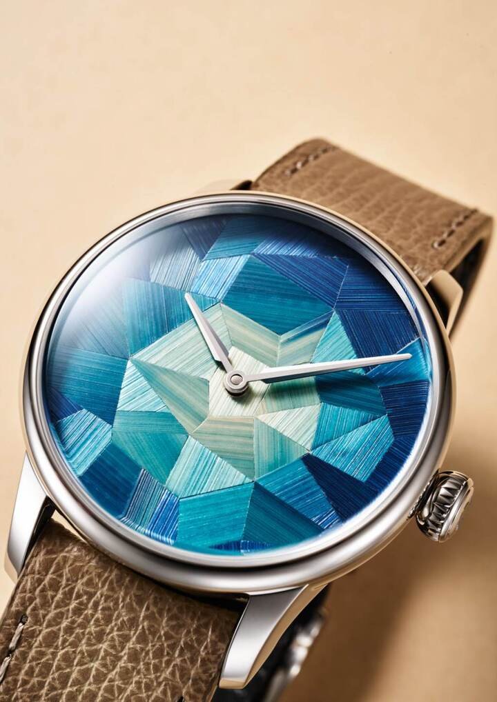 Louis Erard Marqueterie de Paille 腕表以染色黑麦秆构成 “Broken Ice 碎冰” 抽象图案，深蓝、青蓝与浅绿层层交错，呈现宛如冰面折射般的光影。看似几何色块的表盘，近看才会发现一道道天然纤维纹理。28 个区块采不同方向排列，让同一抹蓝色在光线下展现丰富层次。