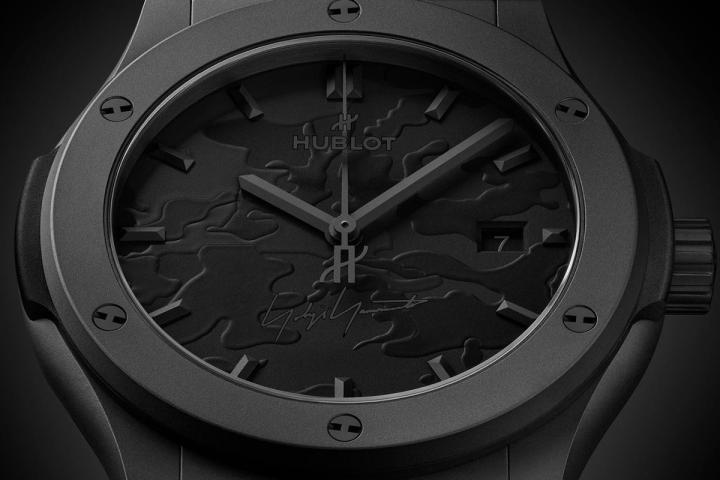 迷彩图案以浮雕层次呈现，透过 "黑上黑" 的设计手法，将不同深浅与质地的黑色叠加于表盘之上。设计师山本耀司的签名低调点缀于 6 点钟位置，成为联名识别的重要细节。
