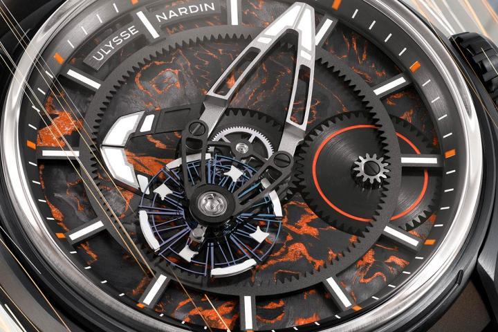 小时盘采用 Carbonium® 碳正离子材料打造，以大马士革钢表面的不规则花纹为灵感，这一材质通过碳纤维与橙色环氧树脂的交织碰撞，呈现极具辨识度的立体效果纹路，令每一枚小时盘皆独一无二。