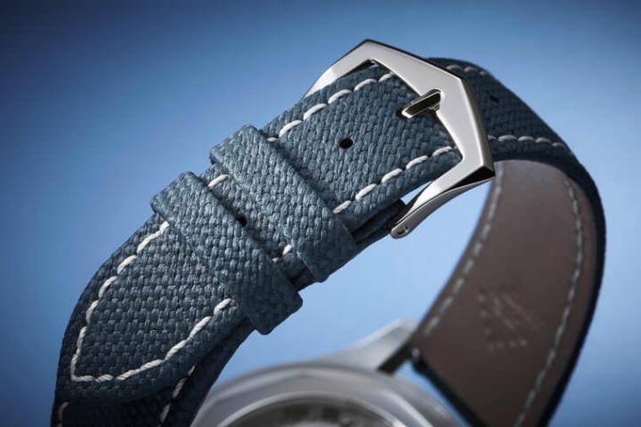 灰蓝色表带为小牛皮材质，由人手缝制并缀以白色缝线，特别之处在于表带表面浮凸有致及展现织物图案，搭配不锈钢针扣